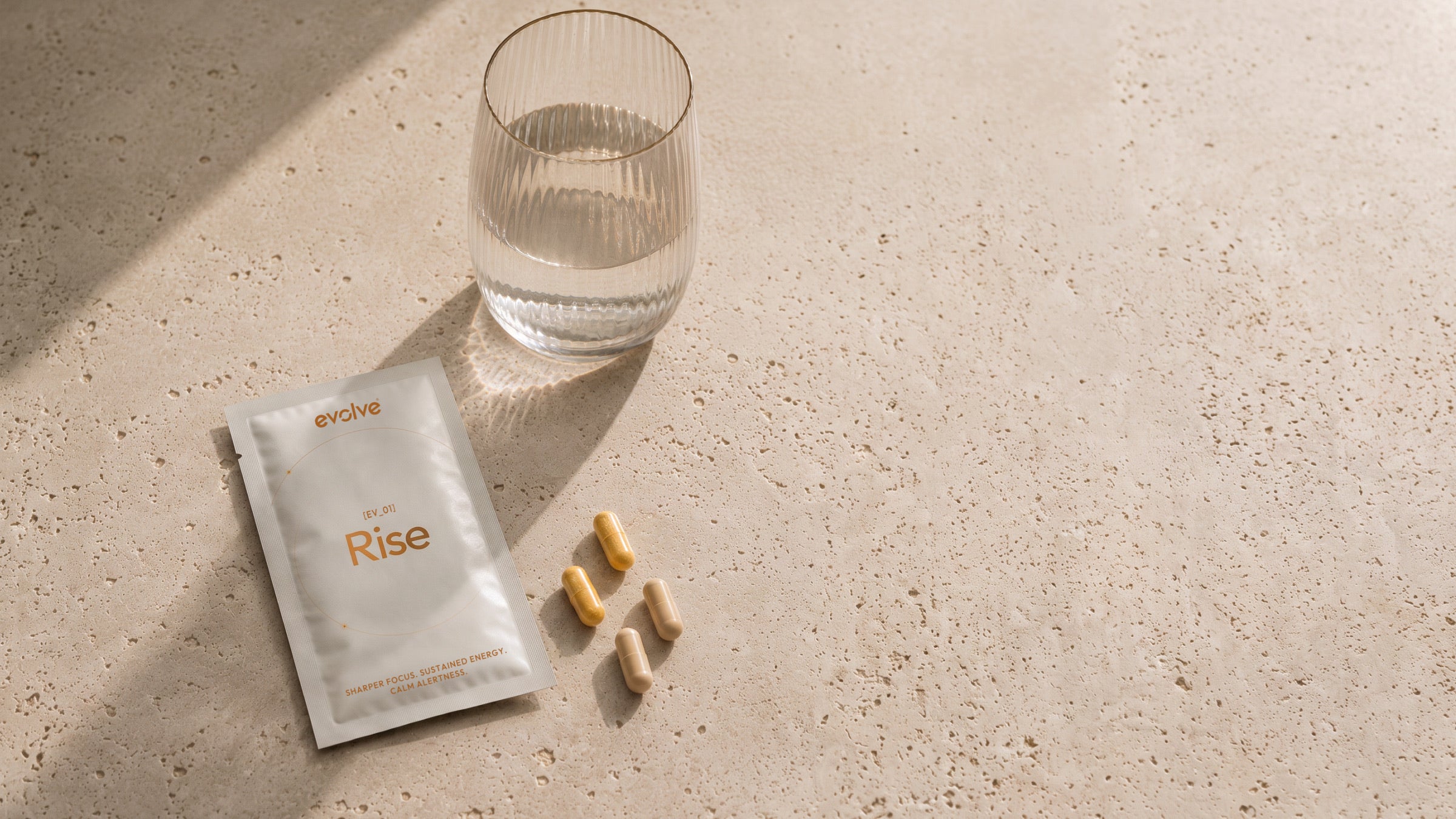 An evolve® Rise sachet, capsules and a glass of water on warm travertine in soft morning light