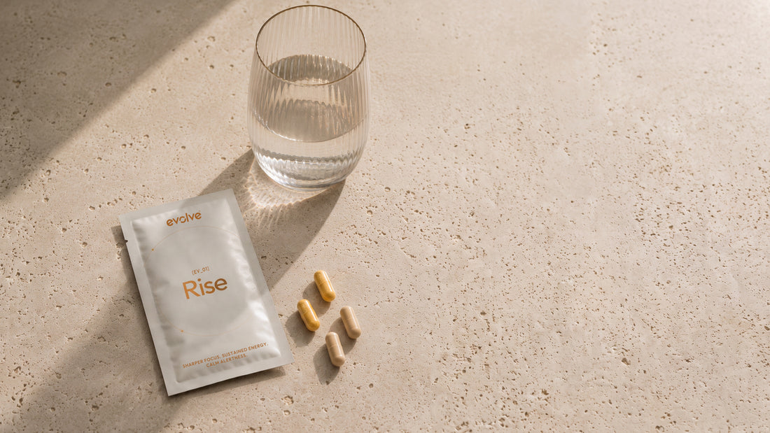 An evolve® Rise sachet, capsules and a glass of water on warm travertine in soft morning light